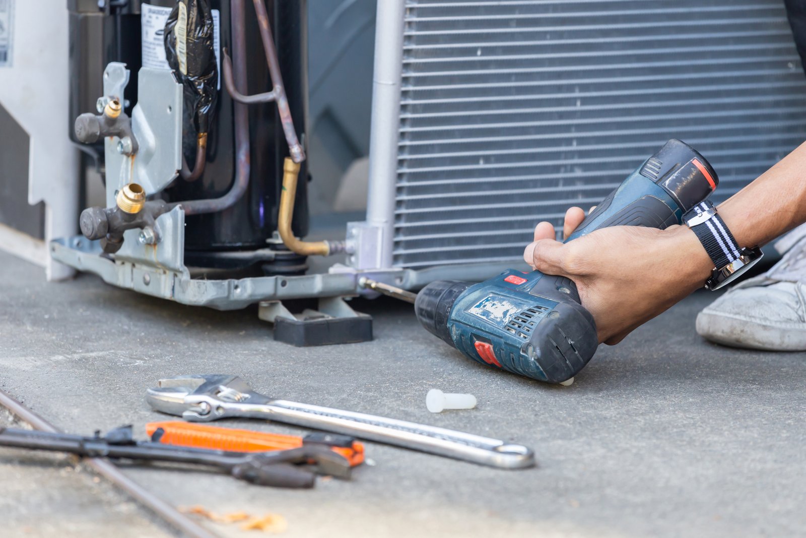 ac repair technician using a power drill to fix an air conditioning compressor unit, with tools scattered nearby for repair work.