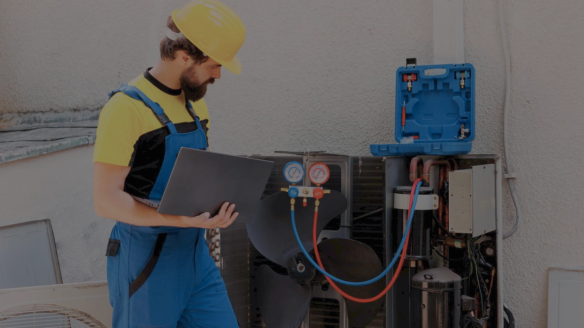 HVAC technician wearing a hard hat and holding a laptop while inspecting an outdoor AC system with gauges and tools